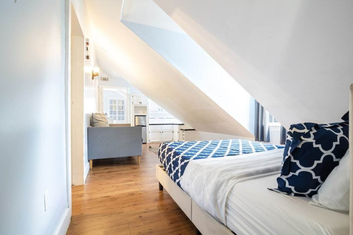 A bedroom with a sloped ceiling, a bed with patterned bedding, a bedside table, and a view of a kitchen area.