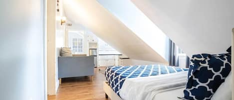 Um quarto com teto inclinado, uma cama com roupa de cama estampada, uma mesa de cabeceira e vista para uma cozinha.