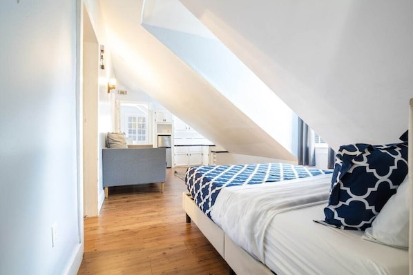 Una camera da letto con soffitto spiovente, un letto con biancheria a motivi, un comodino e vista su un angolo cucina.