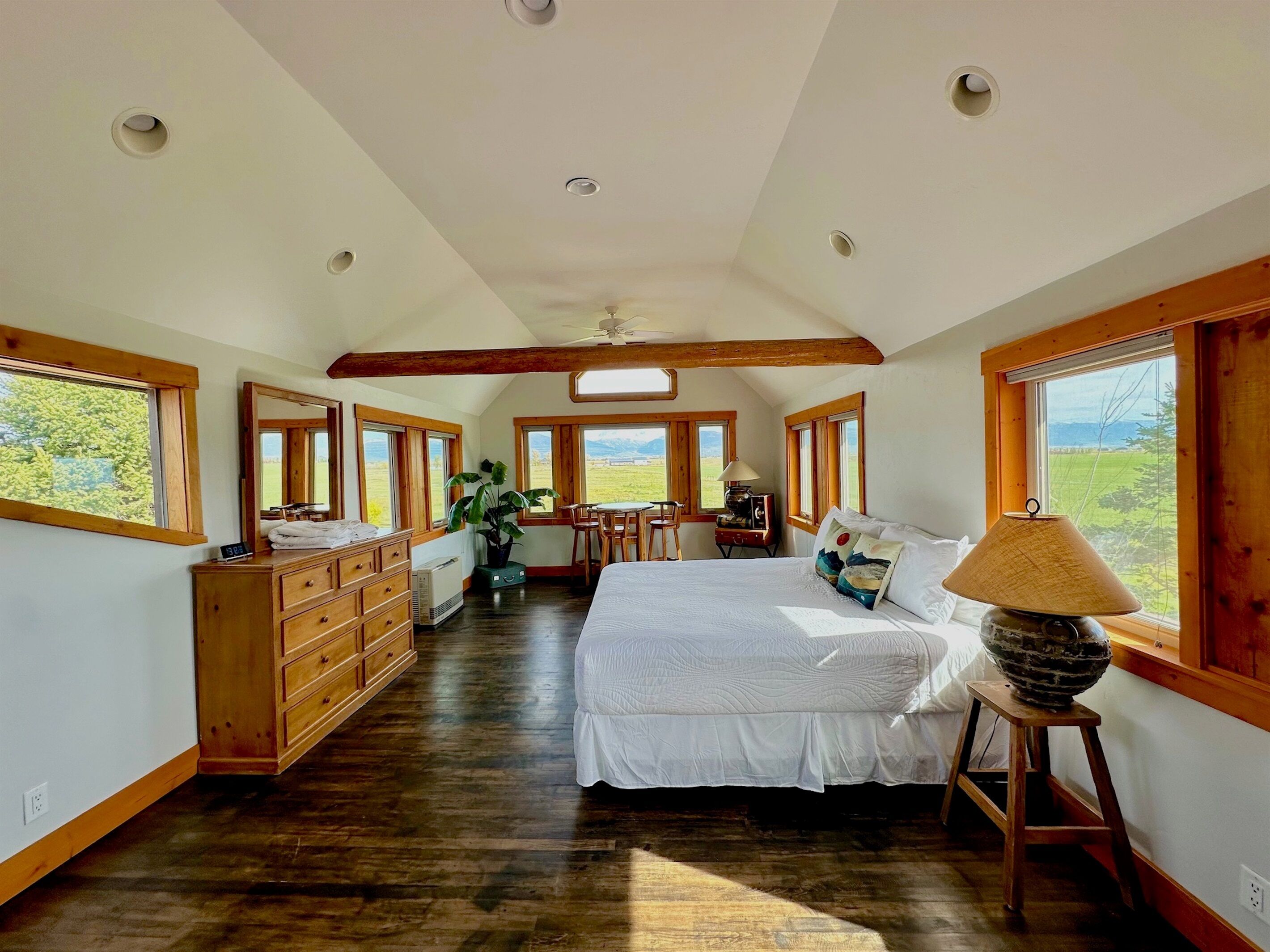 A spacious bedroom with a large bed, wooden dresser, and a desk with a chair. There are multiple windows providing views of the outdoors.