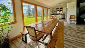 A spacious dining area with a long wooden table and benches, large windows offering a view of a green landscape, and a kitchen in the background. - Teton View - Cache Vista Ranch Estate - Sleeps 32 (Tetonia)