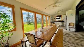 A spacious dining area with a long wooden table and benches, large windows offering a view of a green landscape, and a kitchen in the background.