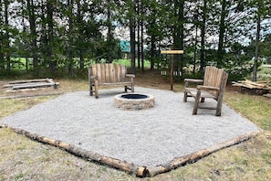 A circular fire pit surrounded by two wooden Adirondack chairs in a natural setting with trees. - Oct Special -Yellowstone Escape -Game Rm -Hot Tub (Island Park)
