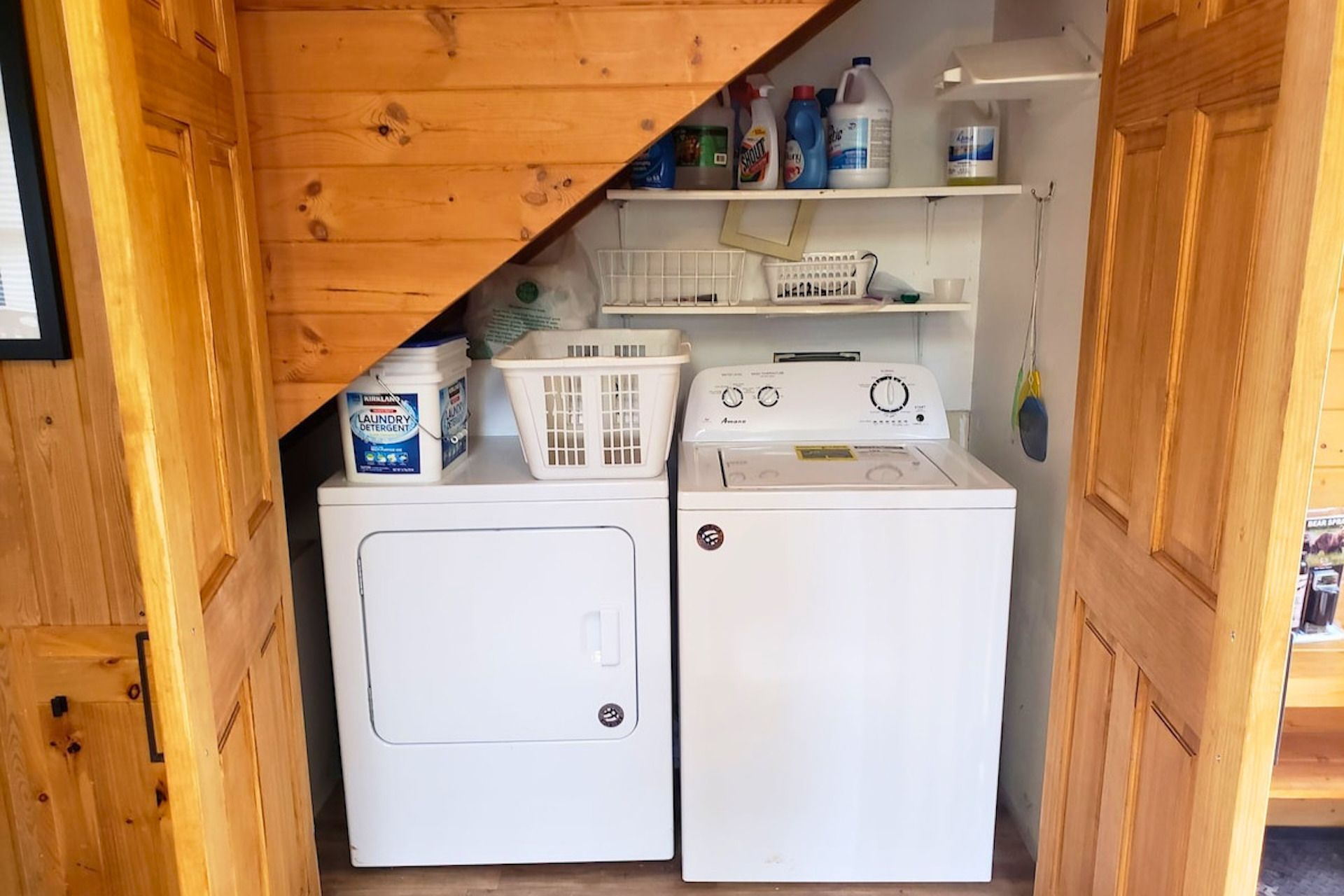 A compact laundry room with a front-loading washer and dryer, a shelf with cleaning supplies, and a basket.