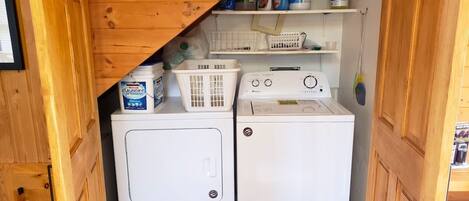 Een kleine wasruimte met een voorlader wasmachine en droger, een plank met schoonmaakmiddelen en een mand.