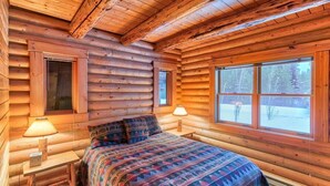 Ett sovrum i en timmerstuga med en säng, två sänglampor och ett fönster med utsikt över träd.