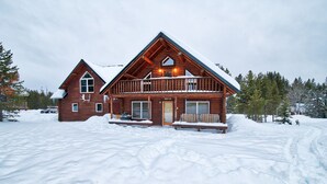 Una cabaña de troncos de dos pisos, con techo nevado, un porche con banca y un camino de acceso visible.