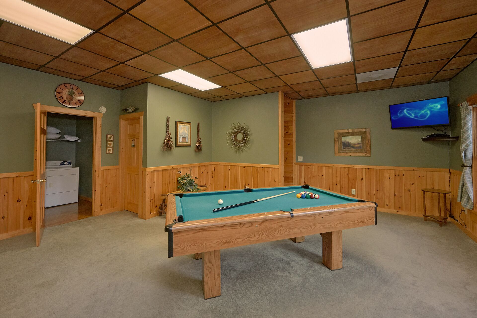 Una mesa de billar en una sala con paneles de madera, un reloj y un televisor en la pared.