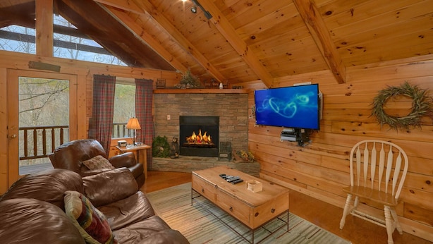 A cozy cabin interior with a leather sofa, a wooden coffee table, a television mounted on the wall, and a stone fireplace. - Affordable Hot tub cabin | Cozy | Quiet (Pigeon Forge)