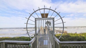 A wooden boardwalk leading to a gate with a circular metal frame and palm tree design, overlooking a beach. - Trendy Beach Condo- Sunset views, Sandy Toes, Surfing more... (1304) (Galveston)