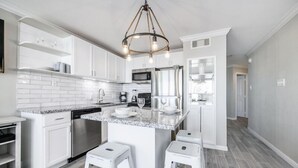 A modern kitchen with white cabinetry, a granite countertop, stainless steel appliances, and a central island with stools. - Trendy Beach Condo- Sunset views, Sandy Toes, Surfing more... (1304) (Galveston)