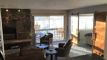 A living room with a stone fireplace, a sofa, a coffee table, and a TV. There is a sliding glass door leading to a balcony with outdoor furniture.