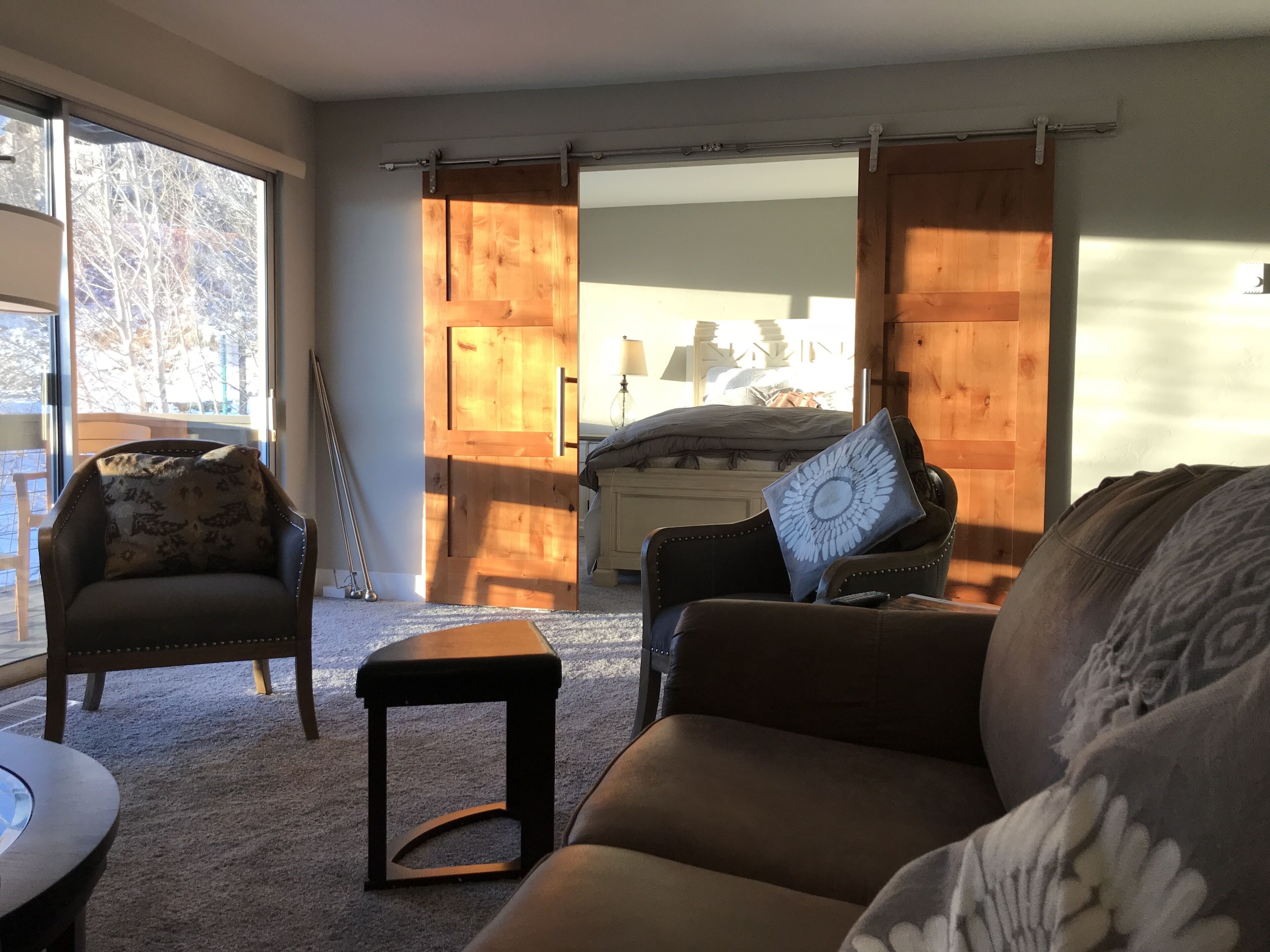 Ein gemütliches Wohnzimmer mit Sofa, Sessel und kleinem Tisch. Eine Schiebetür führt zu einem Schlafzimmer mit Bett und Nachttisch.