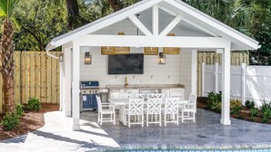 A covered outdoor kitchen with a pool, white furniture, and a TV. - Indigo Cottage: Stylish Island Retreat | Walkable to Everything | Pool + Spa | Dogs Welcome (Saint Simons Island)