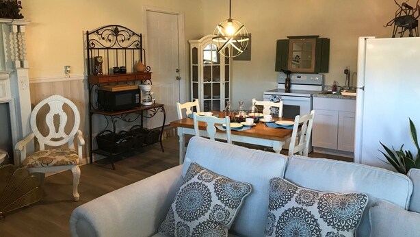 A living room with a sofa, dining table, and chairs. There is a kitchen area with a refrigerator, stove, and cabinets. A chandelier hangs from the ceiling.