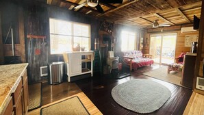 A cozy cabin interior with a wooden ceiling, a ceiling fan, a dining area with a table and chairs, a sofa, and a large window providing natural light.