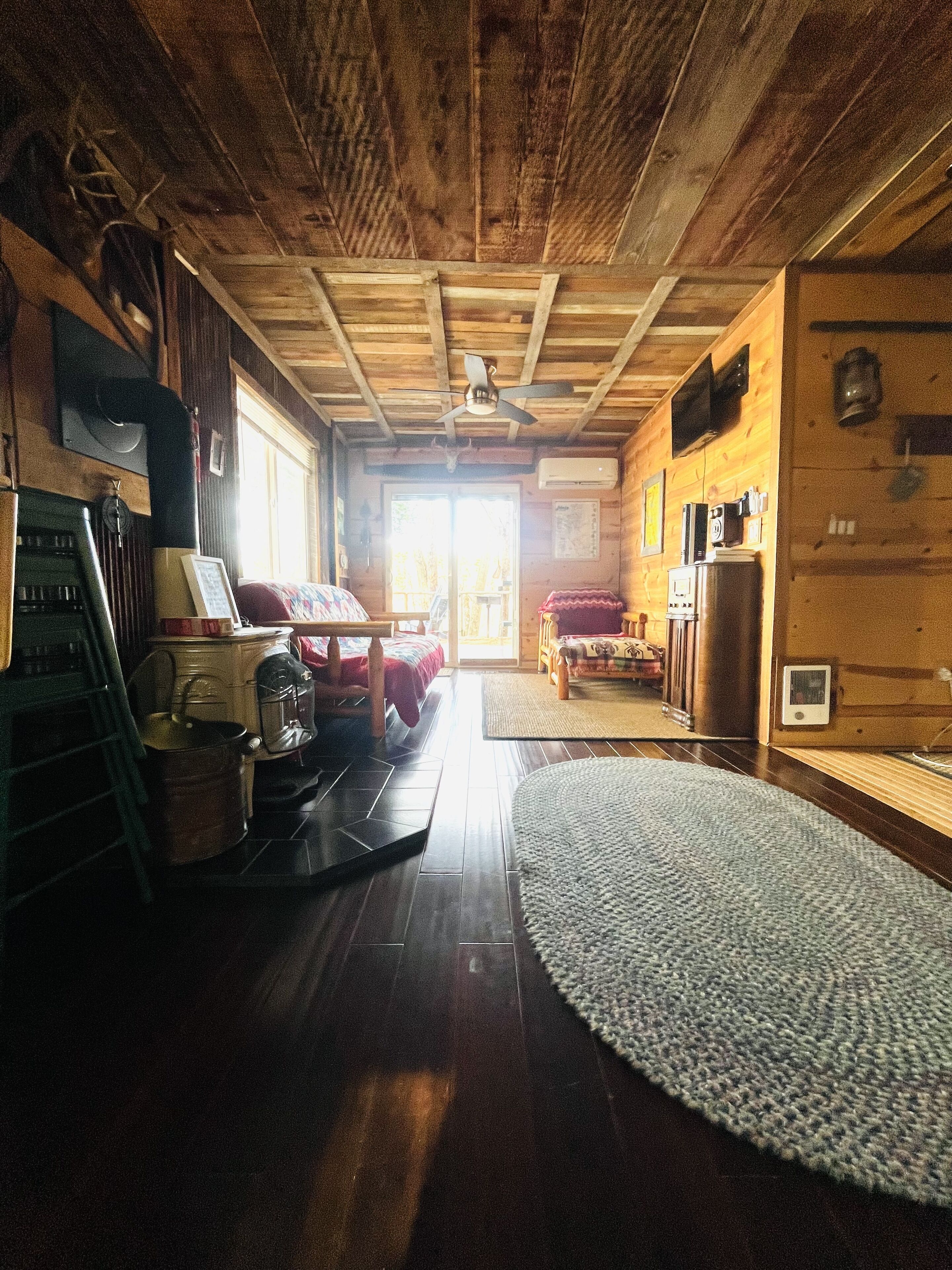 A cozy cabin interior with a wooden ceiling, a ceiling fan, a kitchen area with appliances, and a large window providing natural light.
