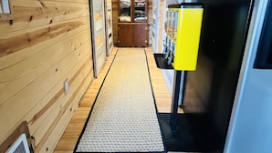A narrow hallway with wooden walls and a central rug leading to a room with a large window and a yellow cabinet.