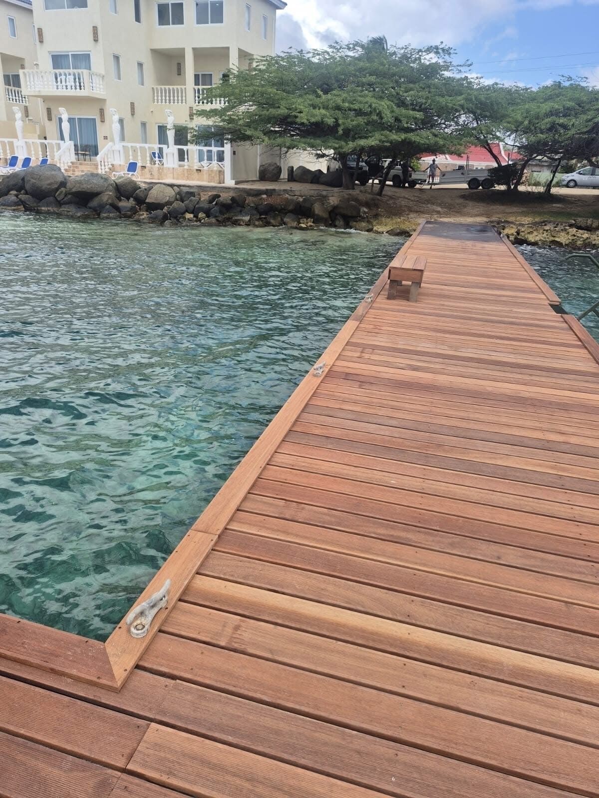 A wooden pier extending into clear water, with a building and trees in the background.