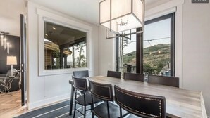 A dining area with a large table and four chairs, a chandelier, and a view of a hilly landscape through large windows. - 3 min walk to Ski/bike main PCMR lift or Main Street!August and September deals! (Park City)