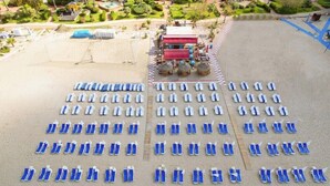 An outdoor seating area with rows of blue lounge chairs and white umbrellas, a red building in the background, and a well-maintained garden. - TAC Premier Hotel - SPA (ALANYA)