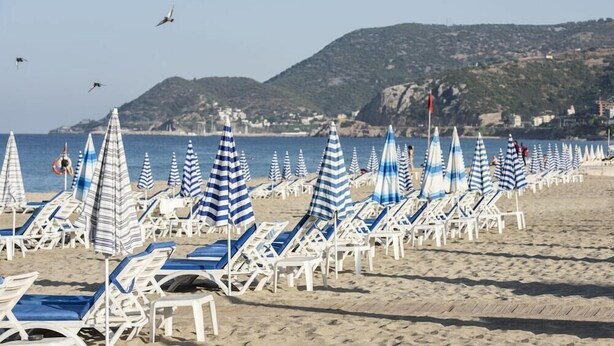 A beach with rows of sun loungers and striped umbrellas, a clear sky, and a hilly landscape in the background. - TAC Premier Hotel - SPA (ALANYA)