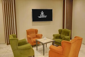 A room with a television displaying 'TAC PREMIER HOTEL & SPA' logo, a green and orange sofa set, and a small table with a glass.