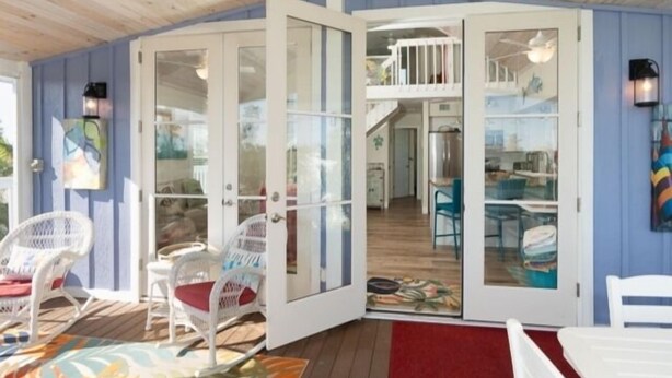 Una habitación con techo de madera, paredes blancas y un ventanal que da a un balcón. Hay sillas y una mesa blancas en la terraza.