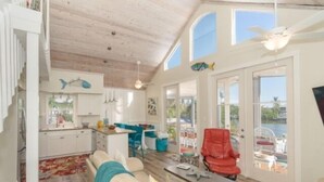 Amplia sala de estar con techo de madera, cocina con alacenas blancas y vista a una masa de agua a través de amplios ventanales.