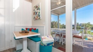 Una habitación con techo de madera, ventana con vista a palmeras, una mesa con cojín y un cesto de basura.