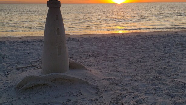 A sand sculpture of a lighthouse on a beach at sunset.
