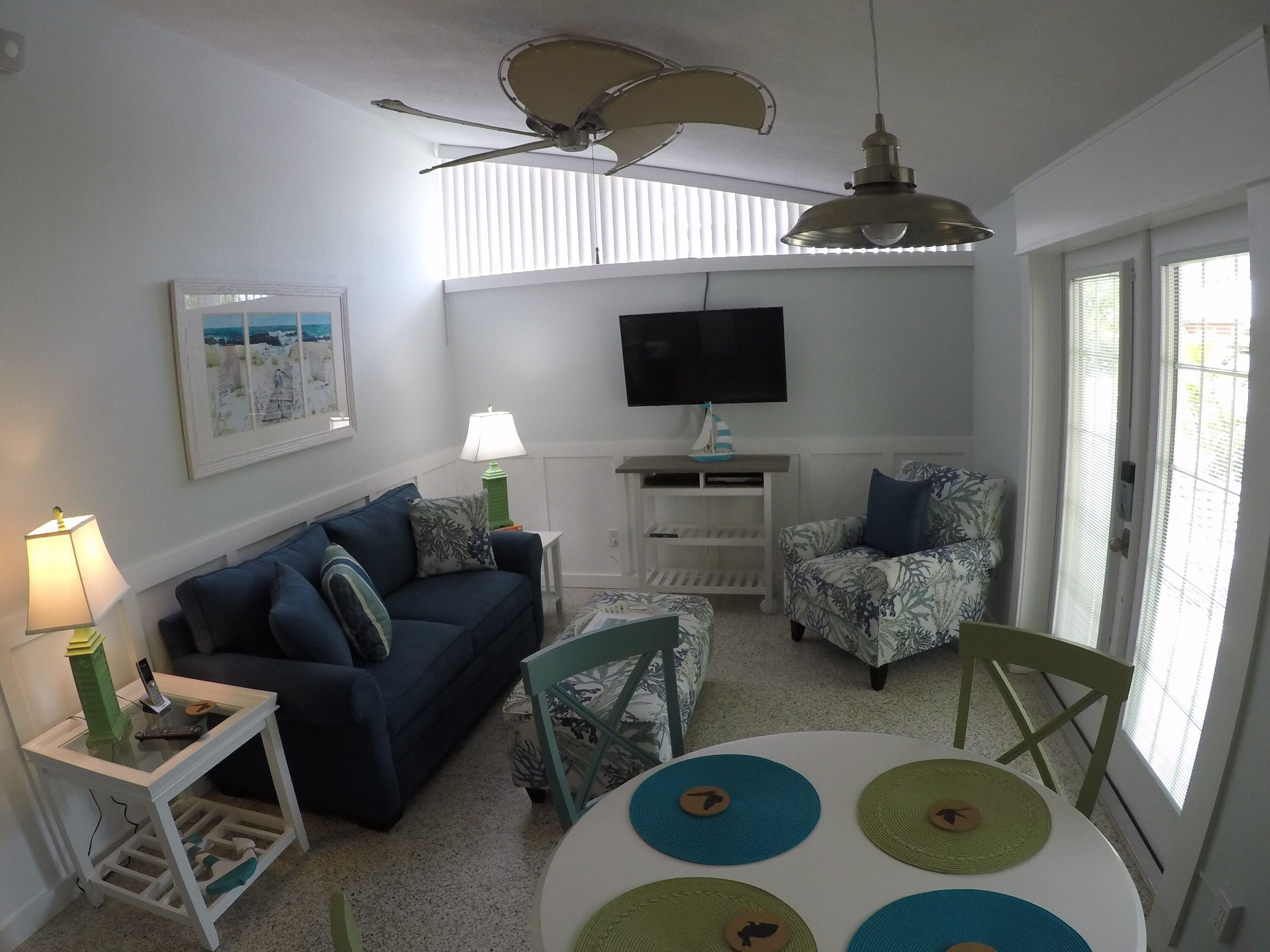 Ein Wohnzimmer mit einem blauen Sofa, einem grünen Sessel, einem runden Tisch mit Platzsets, einem Fernseher und einem Deckenventilator.