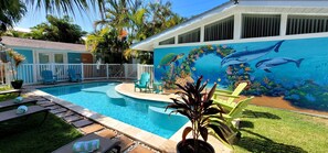 A swimming pool with a mural of marine life, surrounded by a white fence, and a house with a covered patio.