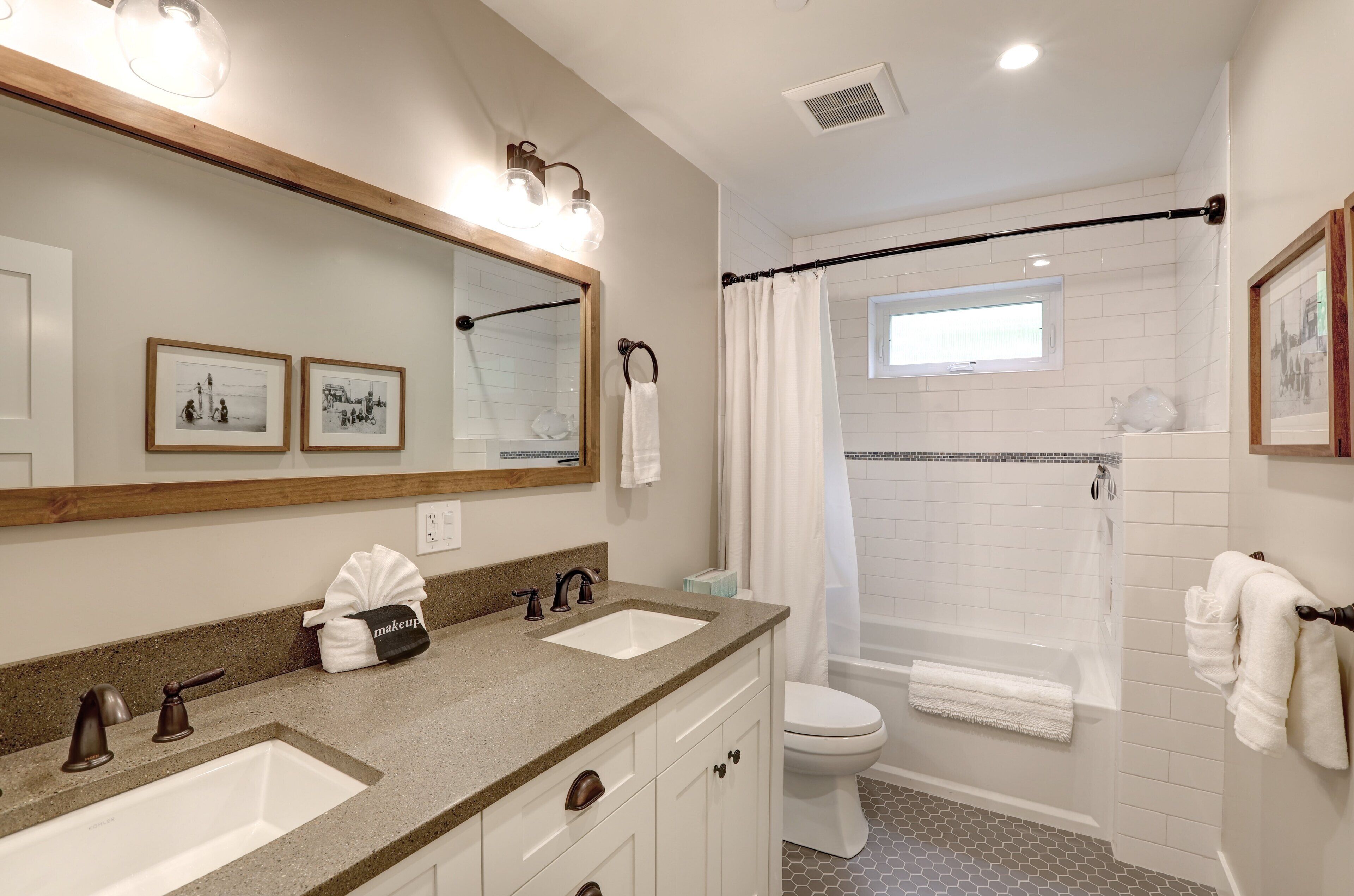Une salle de bain avec deux lavabos, des toilettes, une douche avec une porte en verre et un grand miroir agrémenté de luminaires fixés au mur.