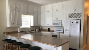 Una cocina con armarios blancos, una isla central con taburetes, microondas, refrigerador y una ventana con persianas.