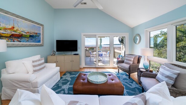 A spacious living room with a ceiling fan, a large painting, a flat-screen TV, and a balcony with outdoor furniture.