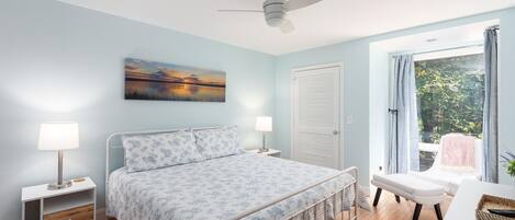Un dormitorio con cama, lámparas de noche, ventilador de techo, ventana con cortinas y vista a la vegetación.
