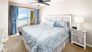 A bedroom with a large bed, a nightstand with a lamp, a ceiling fan, and a view of the beach through a sliding door. - Gulf Dunes 413 • Amazing 5th Floor Condo, Direct Ocean View, Okaloosa Island, Free Beach Service (Fort Walton Beach)