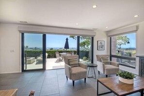 A spacious living area with a large sliding glass door leading to an outdoor patio. There are two armchairs and a small round table with a cushion. A wooden coffee table is in the foreground, and a potted plant is on the table.