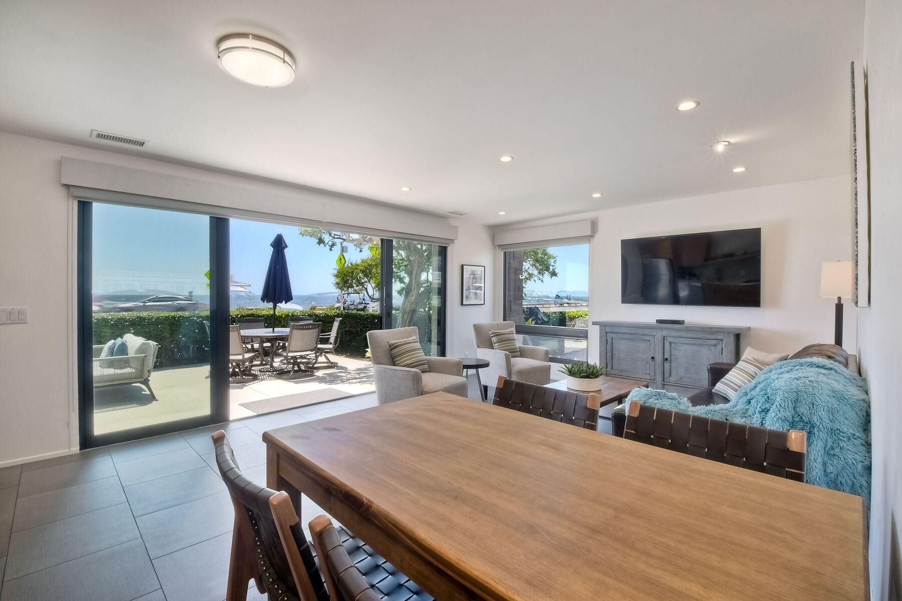 A modern living room with a wooden dining table, comfortable seating, a flat-screen TV, and a large sliding glass door leading to an outdoor patio.