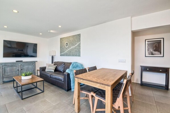 A modern living room with a sofa, a wooden dining table, and a TV mounted on the wall.