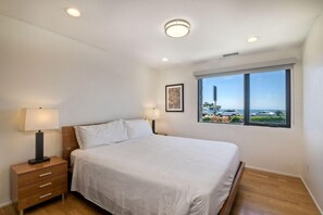 Um quarto com cama, luminárias de cabeceira, mesa de cabeceira de madeira, janela com vista para carros e vegetação, e iluminação embutida no teto.