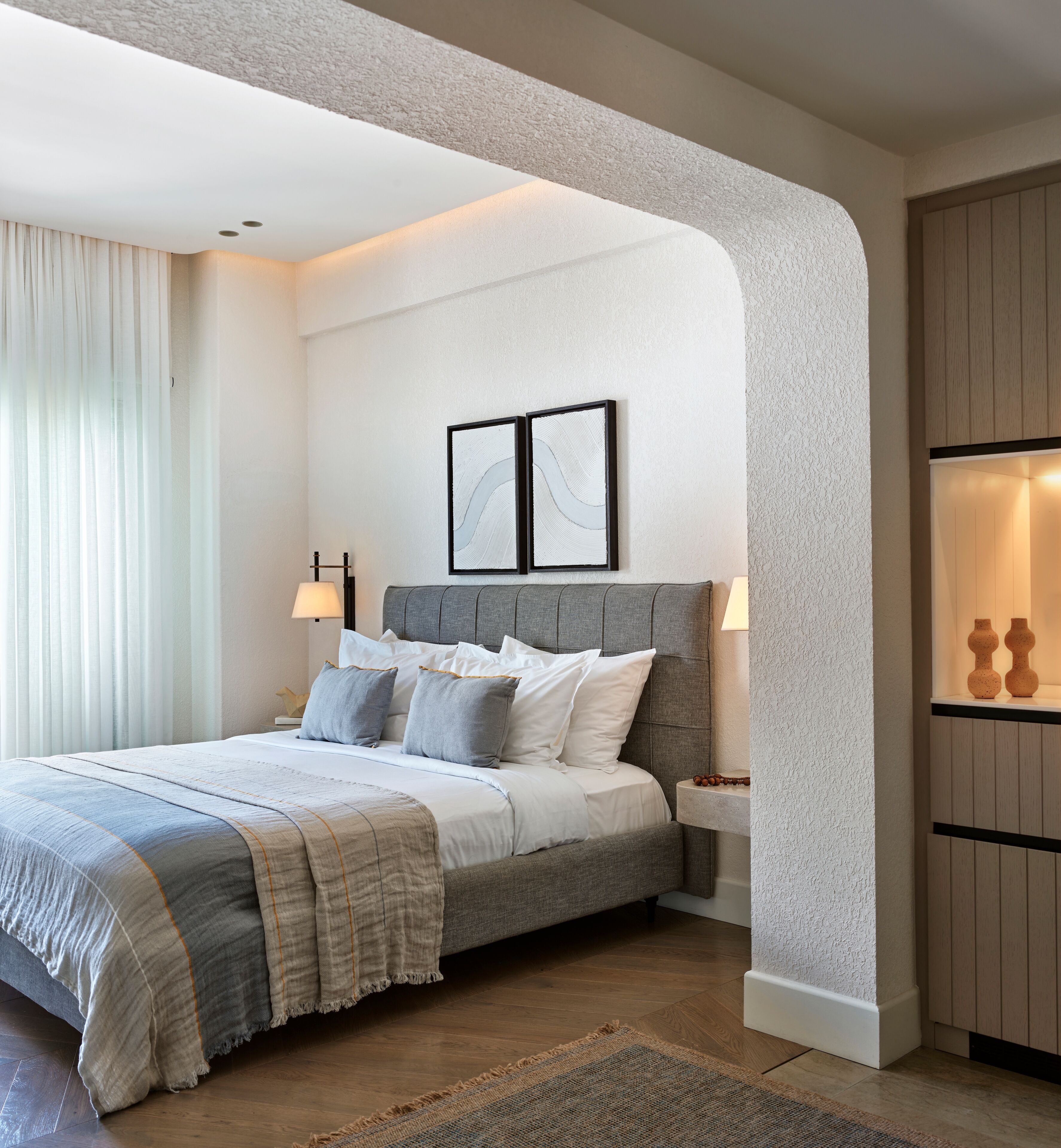 A modern bedroom with a grey upholstered headboard, a large bed with multiple pillows, two bedside lamps, and a framed artwork above the bed.