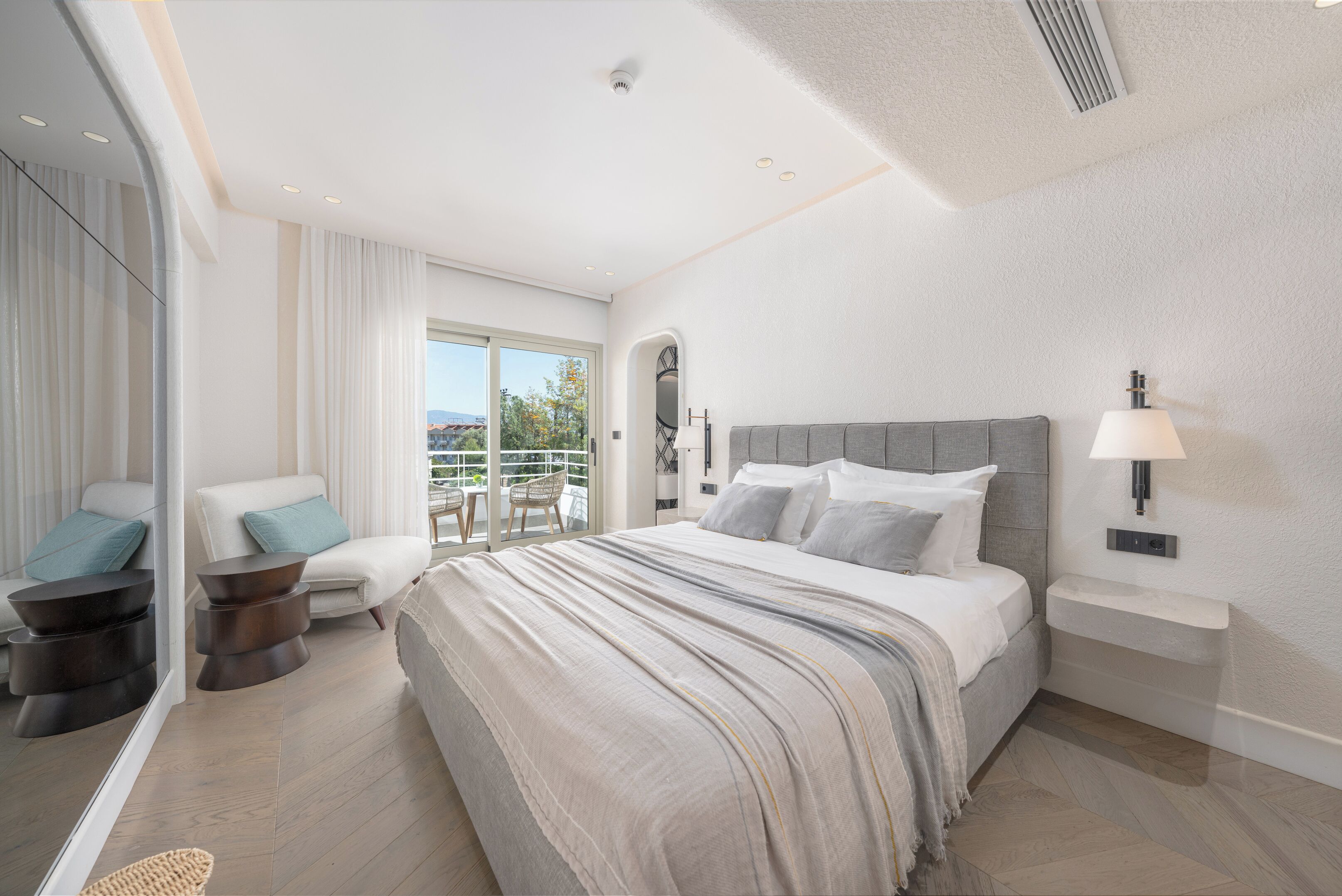 A modern hotel room with a large bed, a seating area with a chair and a small table, a balcony with a view, and a bathroom with a sink and mirror.
