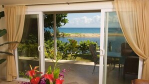 Una habitación con puerta de cristal corredera que da a vista de playa, un área de comedor con sillas y una mesa con un florero.