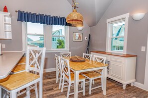 Un coin repas avec une table en bois et des chaises blanches, un meuble blanc et une fenêtre donnant sur des maisons.