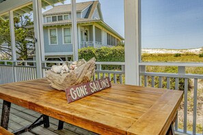 Une table en bois sur laquelle est posée une pancarte indiquant « JE SUIS PARTI(E) VOILER », sur une véranda d’où l’on peut admirer une maison et une plage.