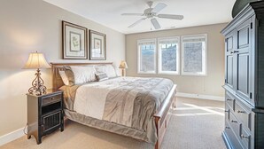 Un dormitorio con cama, mesita de noche, lámpara, ventilador de techo, cómoda y dos ventanas con vistas.