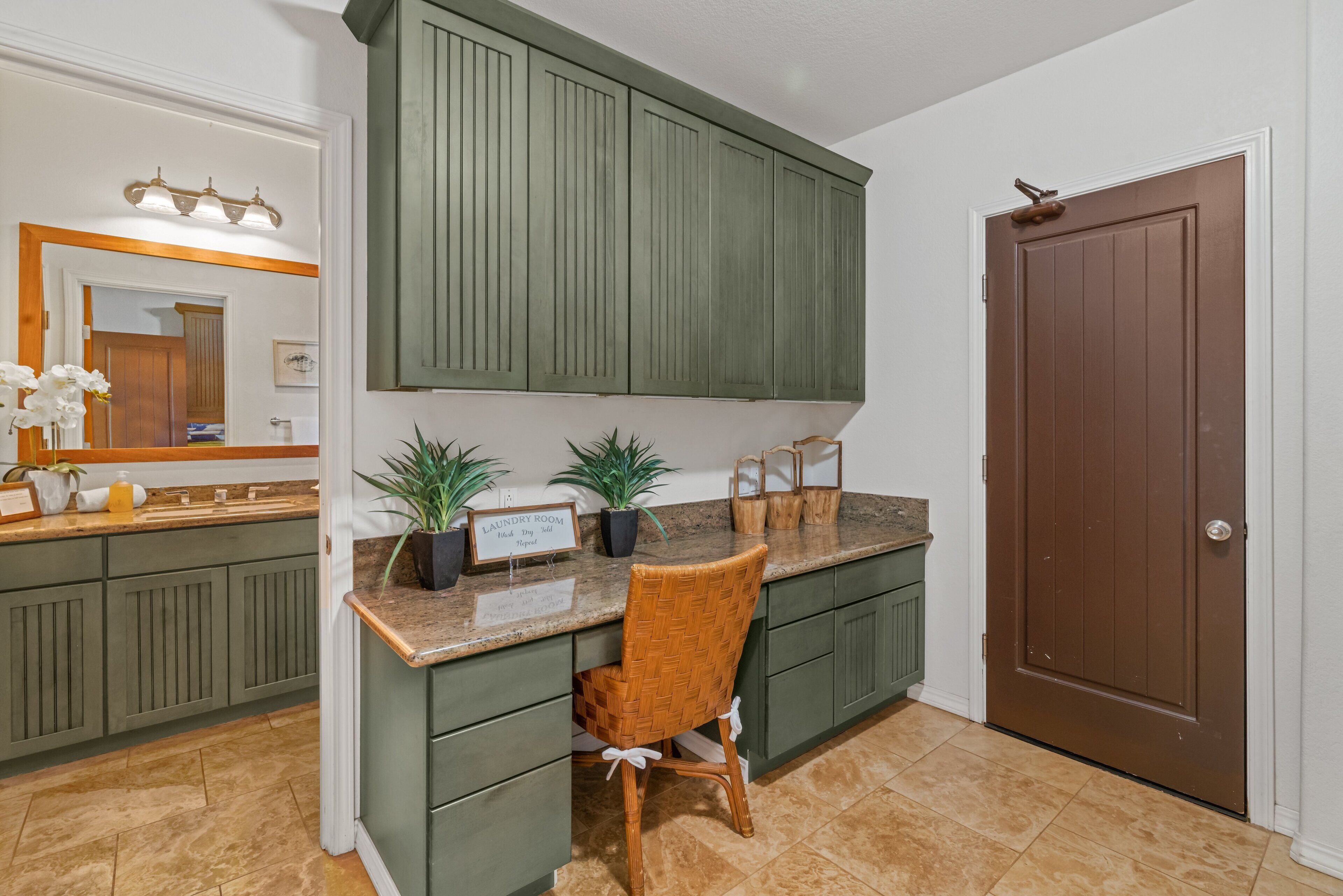 Une cuisine avec des meubles de rangement verts, une chaise en bois, un plan de travail orné de plantes et une porte.
