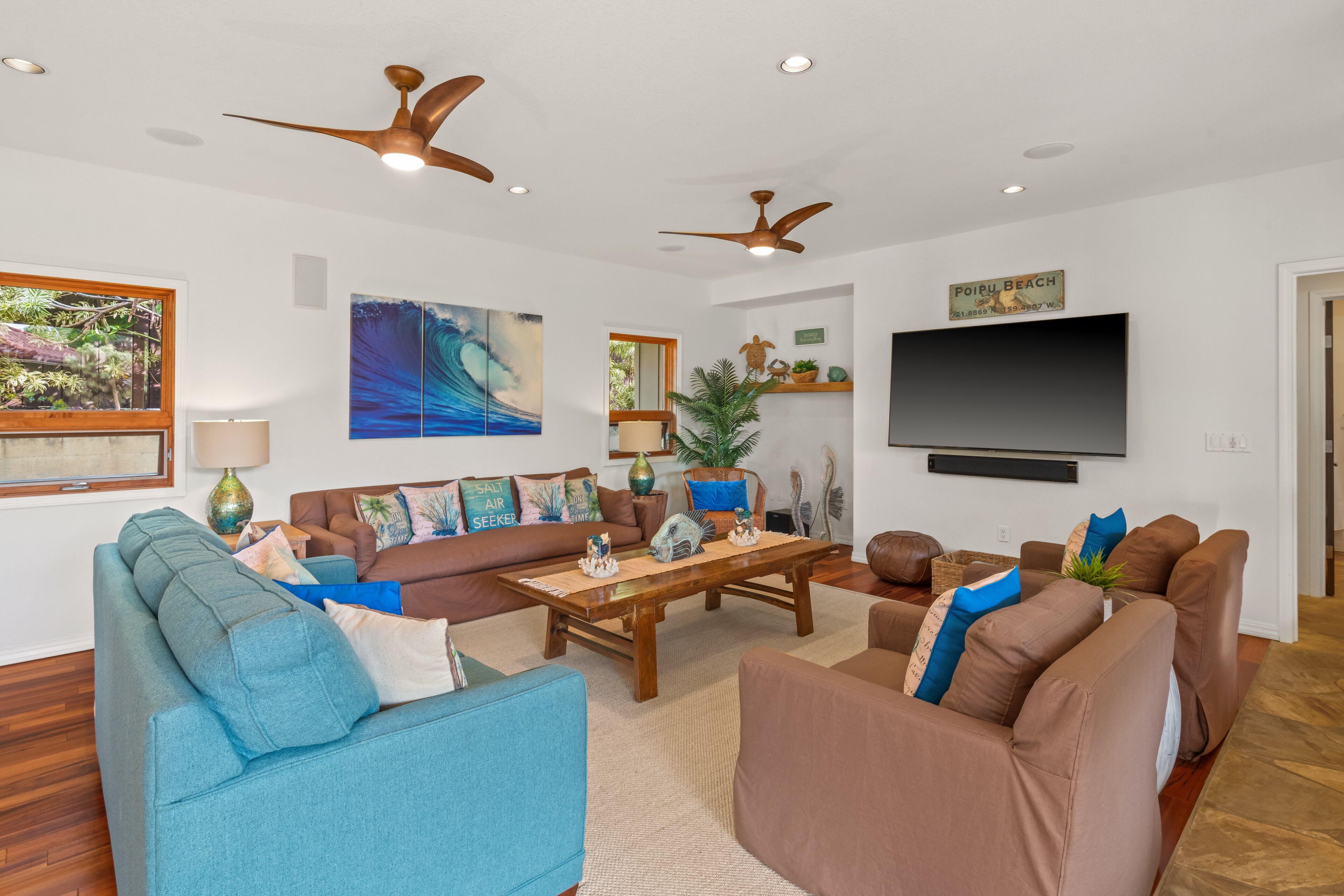 A spacious living room with a large flat-screen TV, multiple sofas, ceiling fans, and a large window with a view of palm trees.